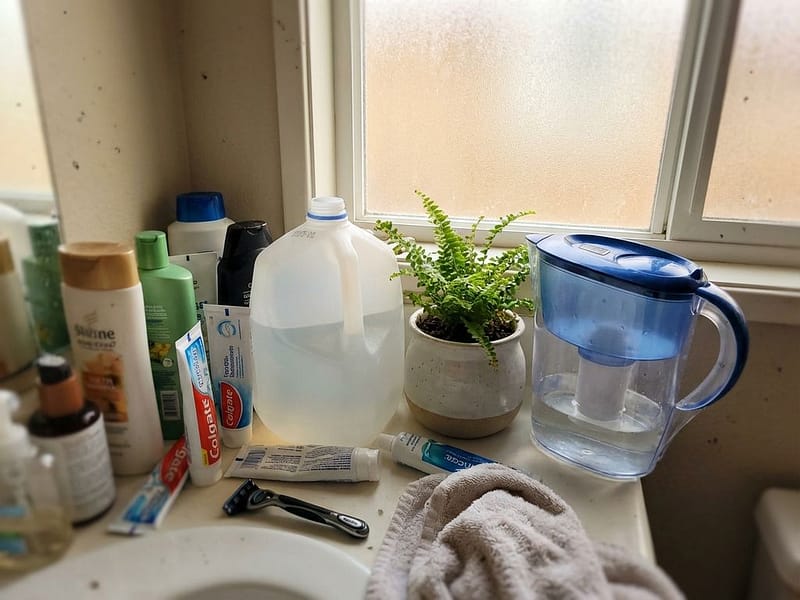 Overvolle badkamertafel met watercontainer, filterkan en toiletartikelen bij raam met ochtendlicht
