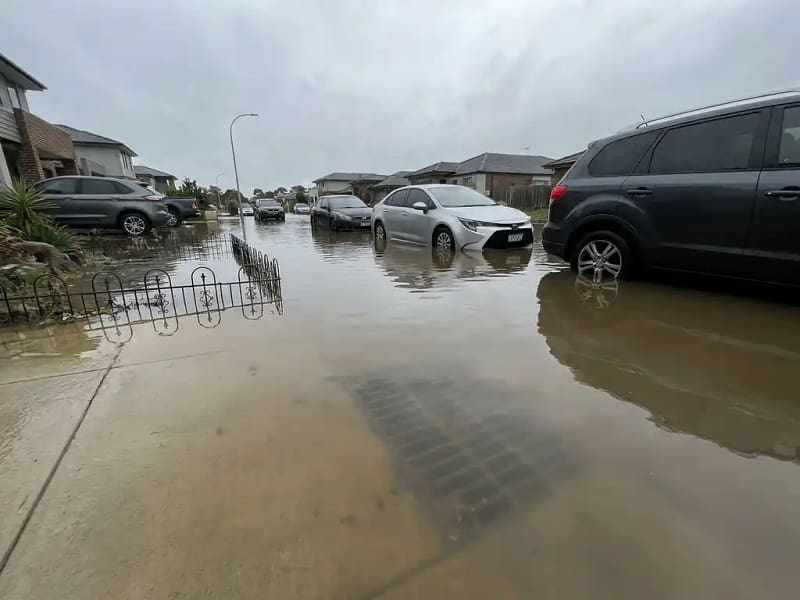 Ondergelopen woonstraat met modderig bruin water rond geparkeerde auto's en tuinhekken, verstopte straatkolk zichtbaar onder het wateroppervlak.