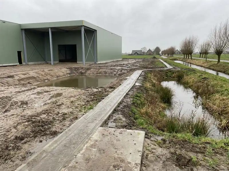 Bouwuitbreiding naast nieuw aangelegde retentievijver in polderlandschap, met regenwater dat via betonnen afvoergoot infiltratiebekken instroomt.