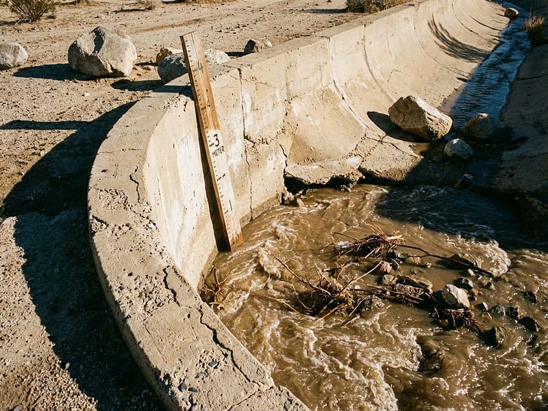 Stromend modderwater door betonnen wadi-kanaal in woestijn met meetstok en puin in de stroom